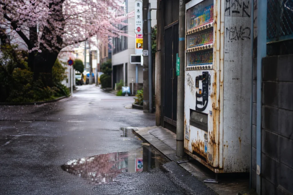 薄曇りの朝、路地裏の水たまりに映る自販機と錆びた金属、濡れた地面の反射